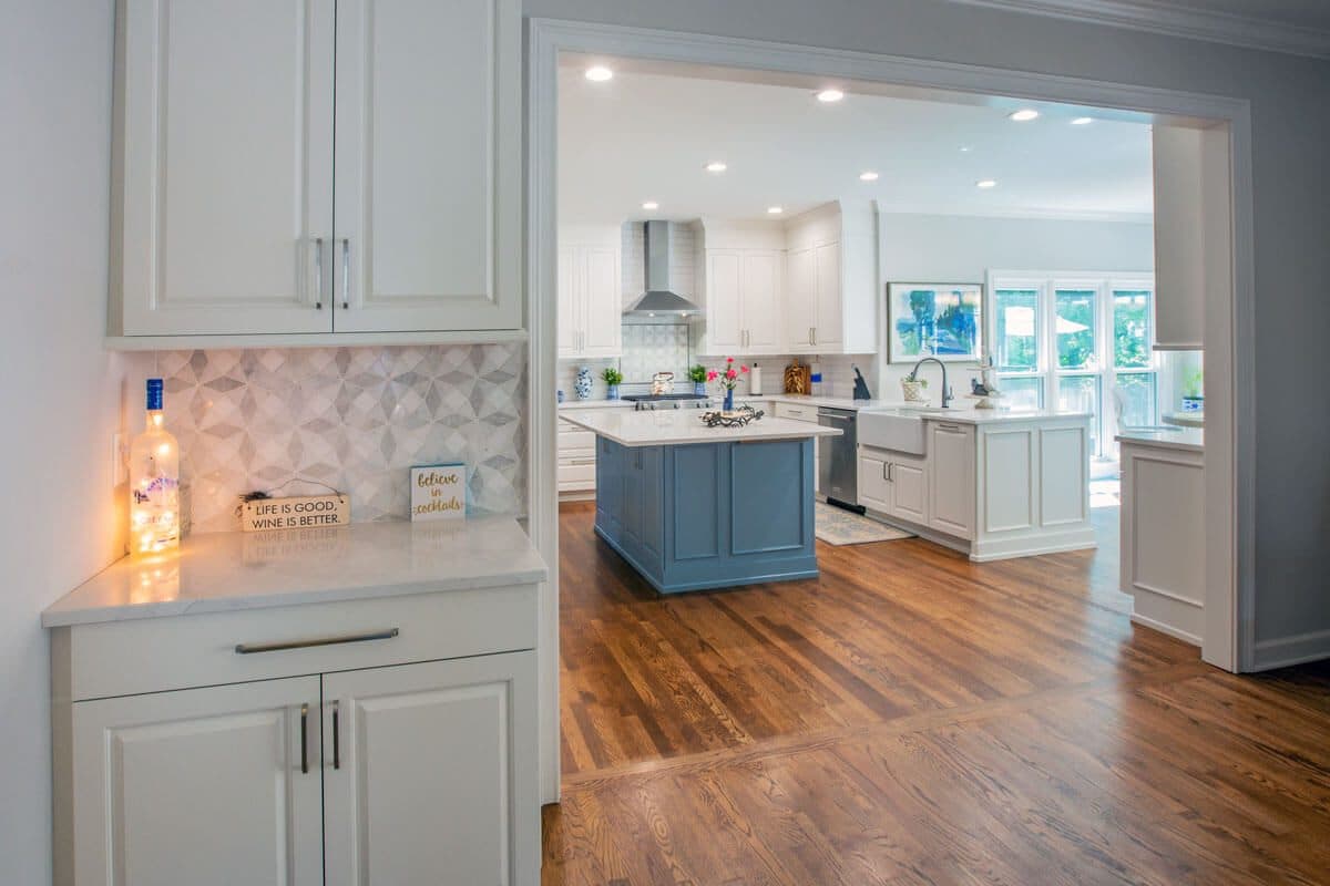 View from a butler's pantry with mosaic backsplash through a cased opening into a white kitchen with a blue island and farmhouse sink
