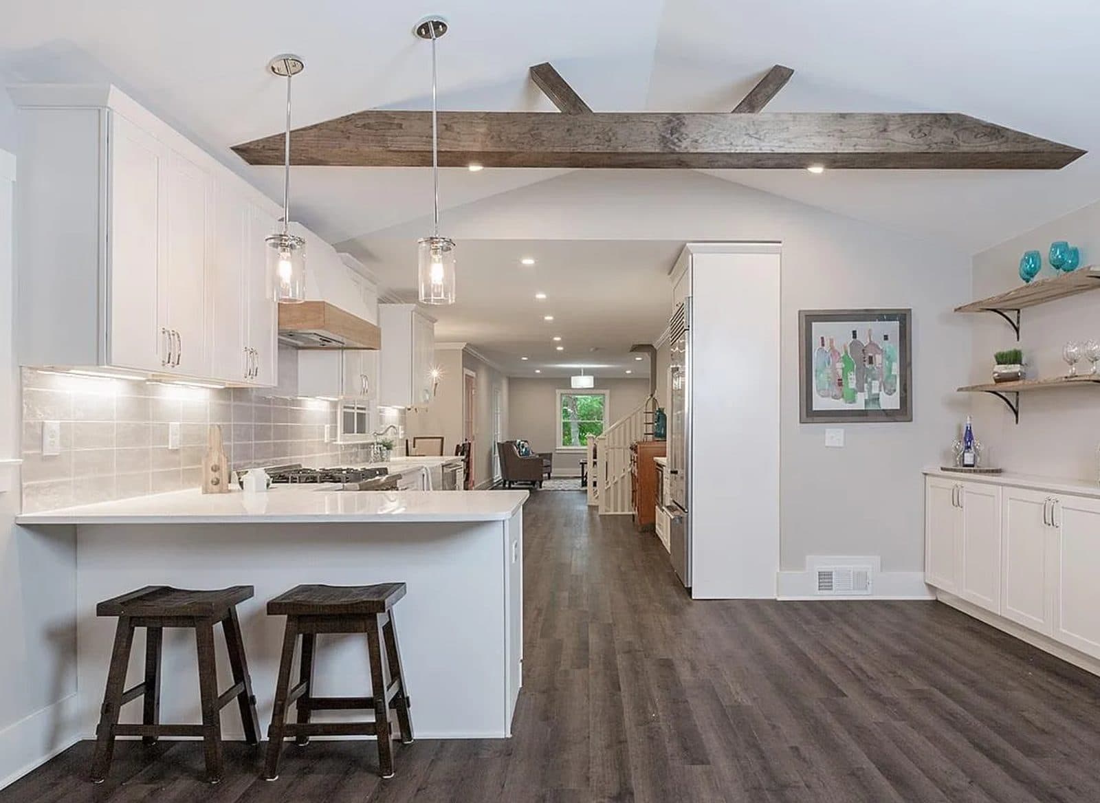Open kitchen with a vaulted ceiling, exposed reclaimed timber beams, white shaker cabinetry, quartz peninsula with bar seating and wide-plank floors
